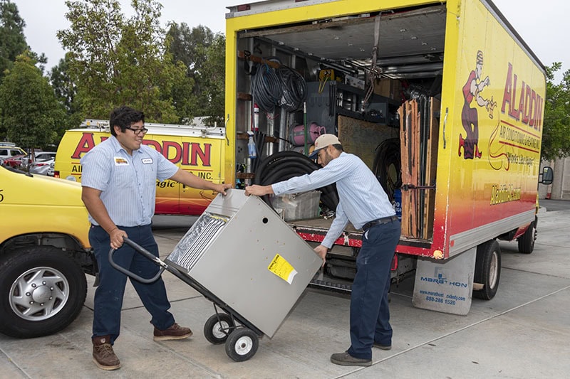 Two HVAC technicians loading an AC unit into a company branded truck.