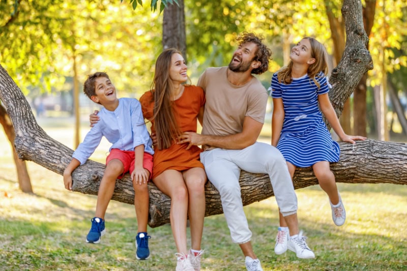 Parents sitting on large tree branch with their children, laughing. Representing four factors to consider when buying a heat pump.